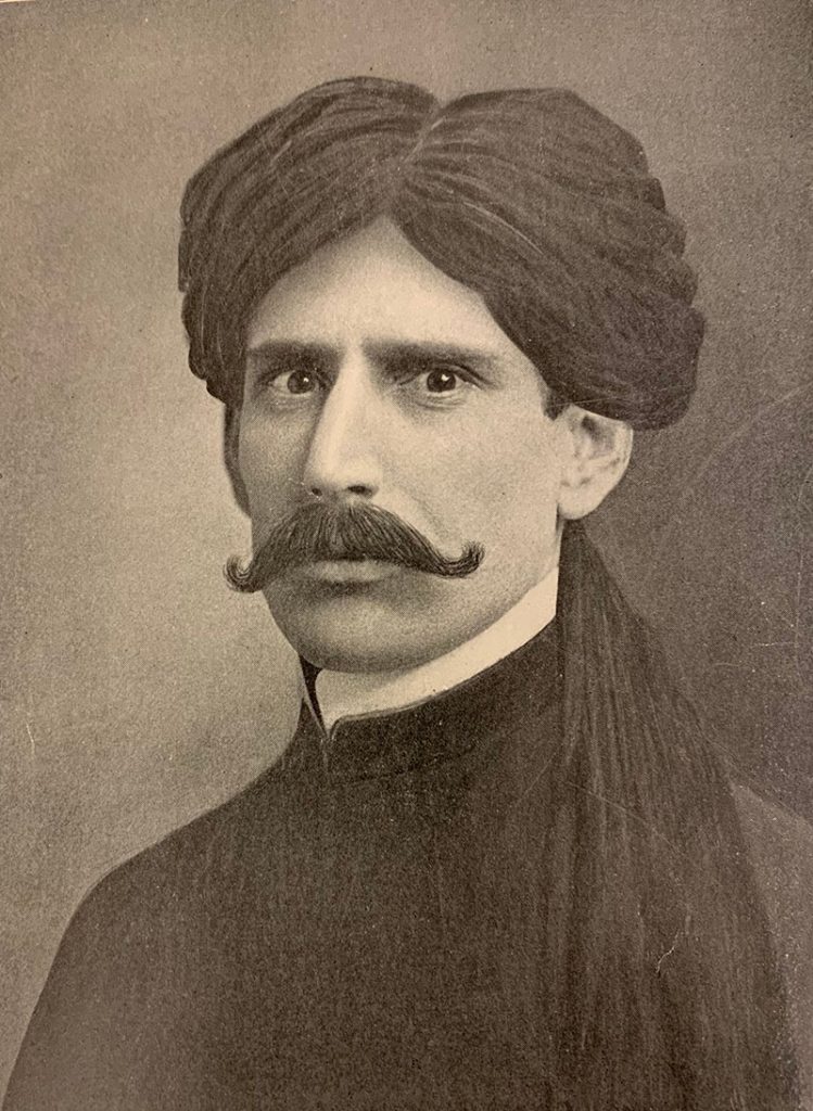 Sepia toned photo portrait of a man wearing a turban, dark eyes looking intently at the camera, and a handlebar mustache, wearing a plain dark jacket with a high collar.