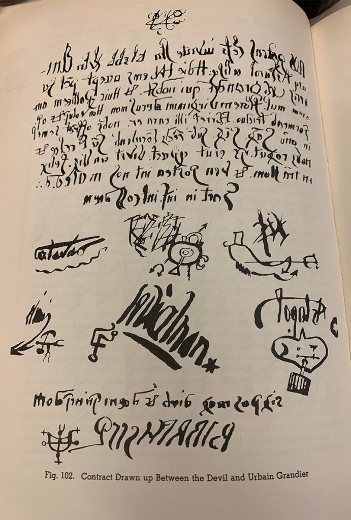 Color photo of a library book page with black script and symbols.  Printed text at the bottom of the page says Fig. 102 Contract drawn up between the devil and Urbain Grandier.