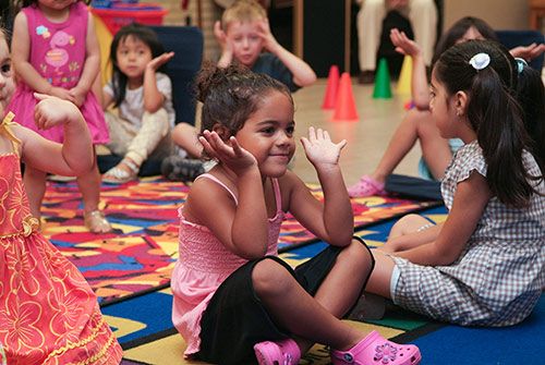 Kids sit together on a play mat and make broad hand gestures.