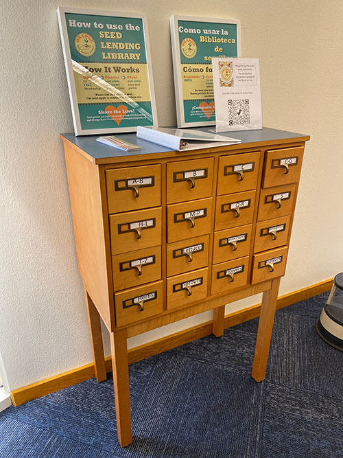 A photograph of Orange Public Library’s Seed Library, re-purposed from a wooden card catalog with multiple drawers, features bilingual signage.