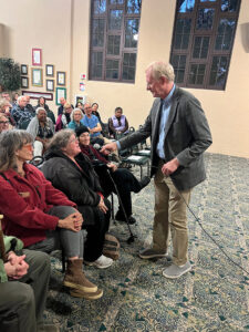 Photograph of Ed Begley Jr. holding a microphone out to an audience member, in a library community room full of seated adult event attendees.