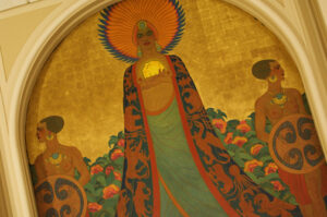 Mural showing three topless Black women, one in center with a large circular crown holding a gold stone in her arms and the others on either side of her holding shields. They are set against a sky made of gold leaf.