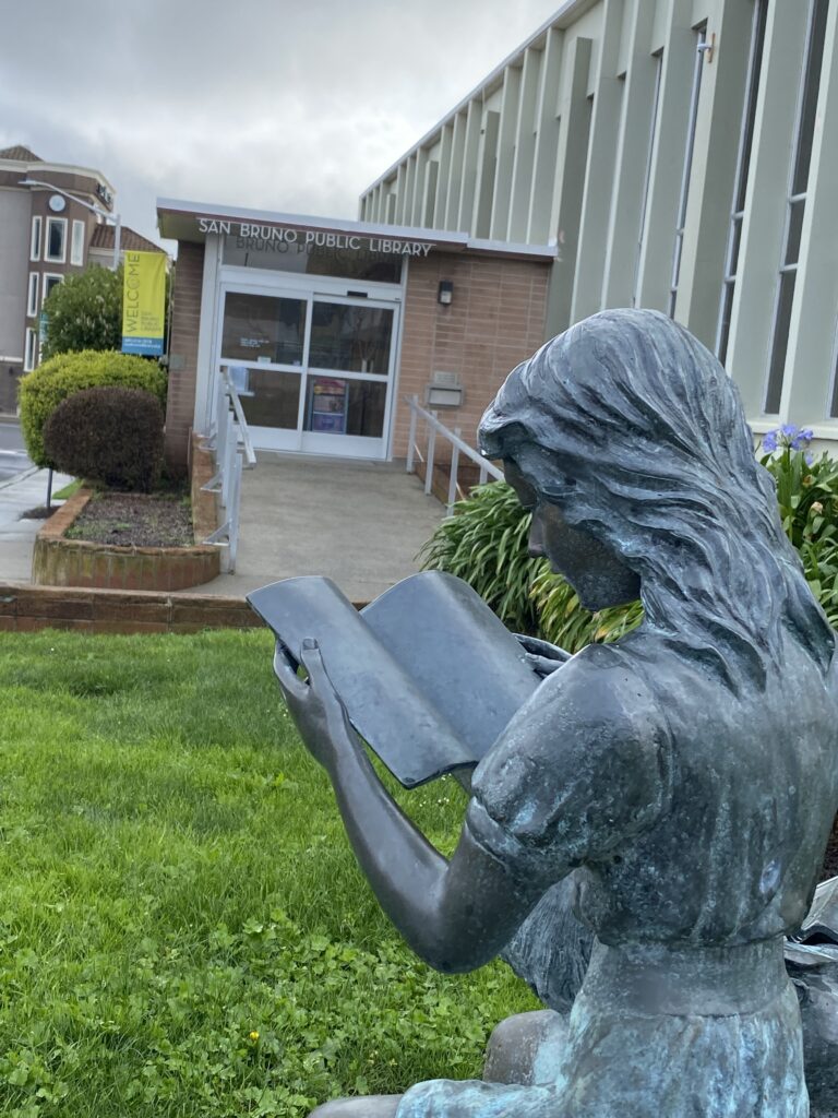 A stone statue of a girl reading in front of a building with a sign that reads "San Bruno Public Library".