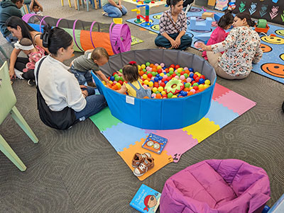 Adults and children engaging with educational manipulatives (small ball pit, crawling tunnel) in the library.