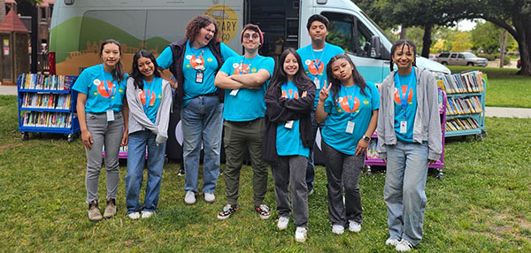 Photo of a group of teens standing in front of a vehicle with cart of books in background.