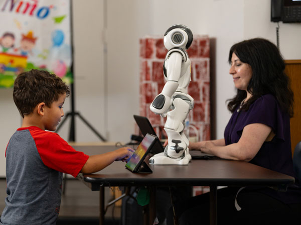 A child interacts with a robot using a touch screen. An adult facilitates behind a laptop. The robot is a NAO robot for social interaction.