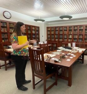 Woman holding a yellow folder stands beside a table with library materials on display.