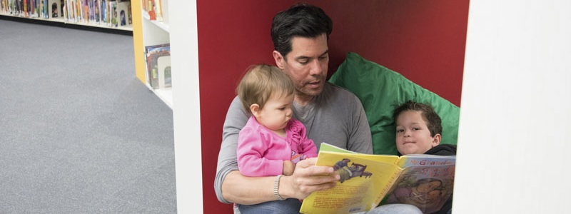 A man reads a book to two children in a comfortable nook at a library.