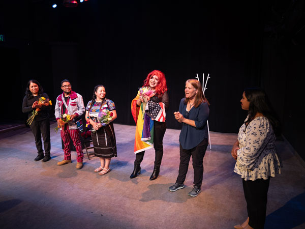 Four performers on stage with bouquets of flowers with several supporters clapping and smiling.