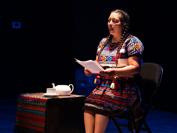 Single female performer seated on a stage in colorful traditional clothes speaks to the audience.