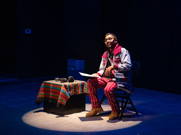 Single male performer seated on a stage speaks to the audience.