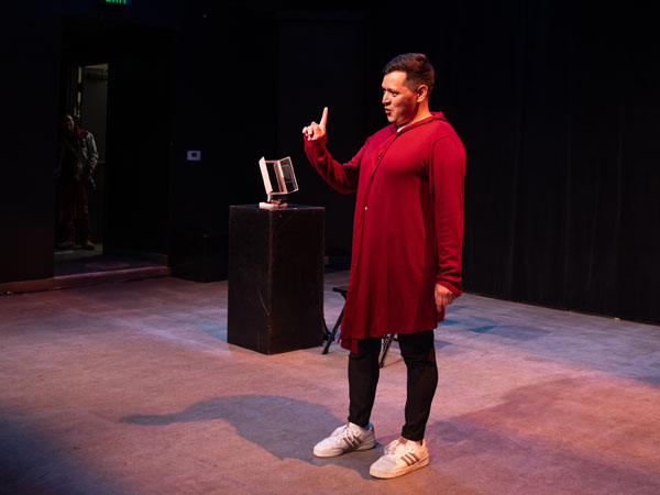 Single performer stands on a stage with their hand held up to the audience pointing with a single finger and their lips are pursed.