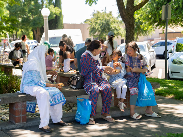 A mother, grandmother, and two daughters sit on a bench outside while the daughters eat lunch.