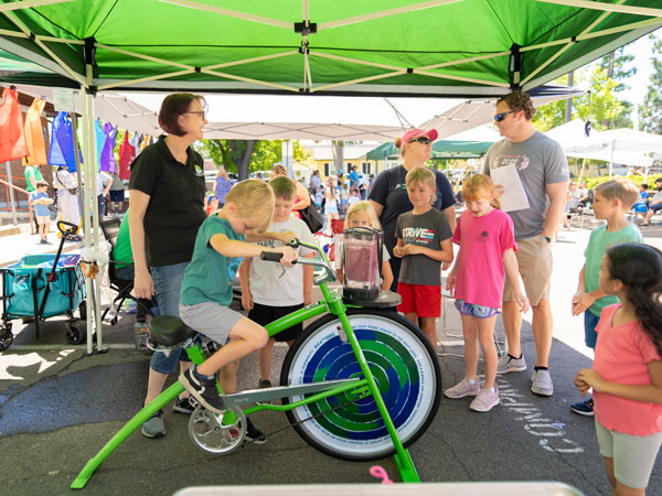 A young child rides a bicycle that powers a blender to make a smoothie while a group of other children and parents watch.