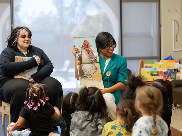 Two library staff workers lead a storytime to a group of young children. The library workerse are sitting on chairs and displaying a picture book to the children.