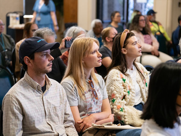 A crowd of seated adults listens to a speaker, not pictured. One audience member holds up a cell phone with the camera facing the speaker.