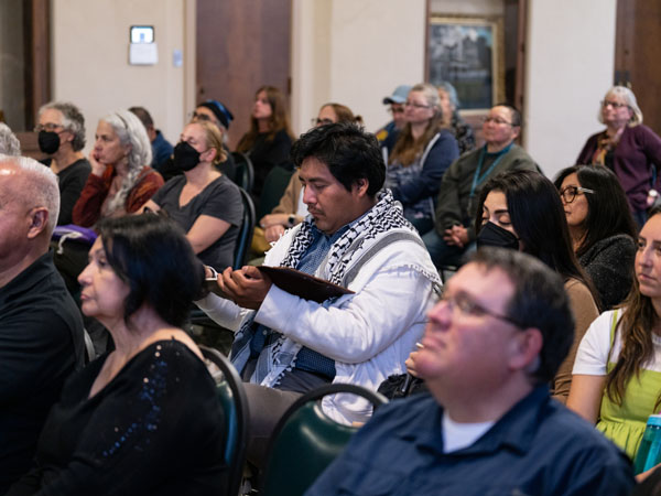 A crowd of seated adults, the focus is on one a man reading a book or taking notes in the center, surrounded by a variety of adults, varying in age.