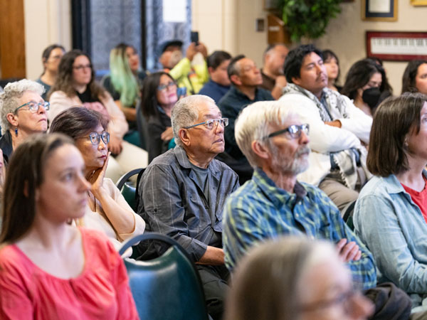 A crowd of seated older adults sits listening to a speaker, not pictured. The focus is on several older adults in the middle of the photo. 