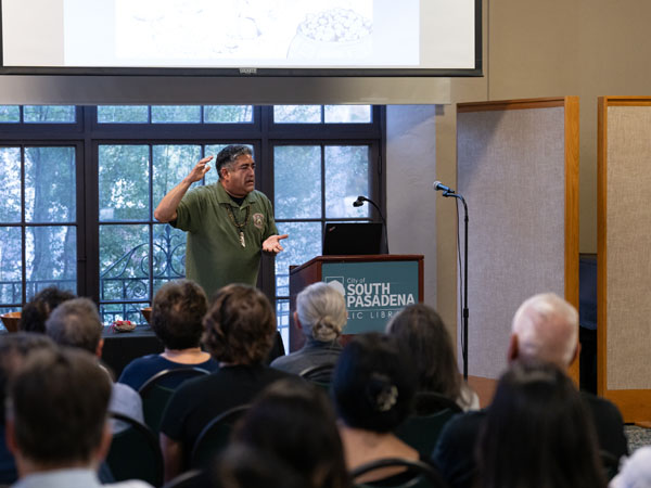 Matthew Teutimez speaks to a crowd of adults. He wears an olive green polo shirt and gestured with his hands. The back of heads of audience members are at the bottom of the image, with the speaker, podium and media screen at top.