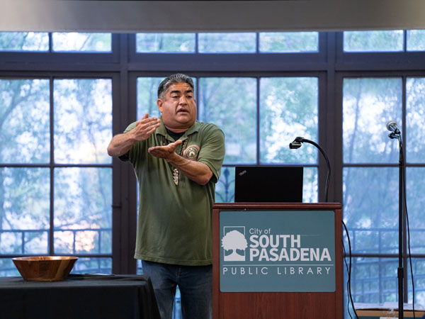 Matthew Teutimez makes a cutting or chopping hand gesture, standing at a podium, with a wooden bowl on a nearby table.