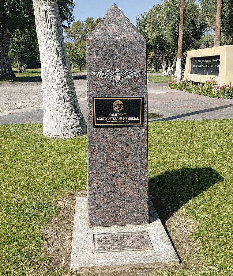 A granite obelisk stands in a parklike setting. Carved into it is an eagle with outstretched wings and a pink triangle on its chest; it holds a bundle of arrows in one talon and an olive branch in the other. Below the eagle, a plaque bearing the Great Seal of the State of California reads, “California LGBTQ Veterans Memorial, dedicated on April 27, 2019.” Another plaque is inset into the foundation of the obelisk and reads, “This memorial is dedicated in honor of gay, lesbian, bisexual and transgender veterans who gave the ultimate sacrifice for the cherished freedom we hold dear and enjoy today. Dedicated May 27, 2001 by AMVETS Post 66.”