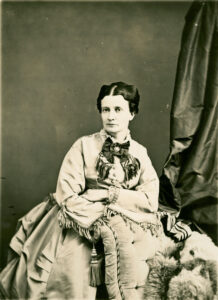 Black and white studio portrait of a middle-aged woman leaning against a chair wearing a dress adorned with a large bow. 