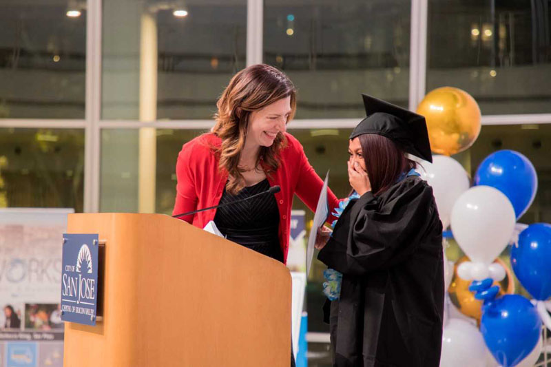 Adult woman congratulating a female in a cap and gown as she walks across the stage at her Career Online High School Graduation. Photo credit San Jose Public Library Marketing Communications Department.