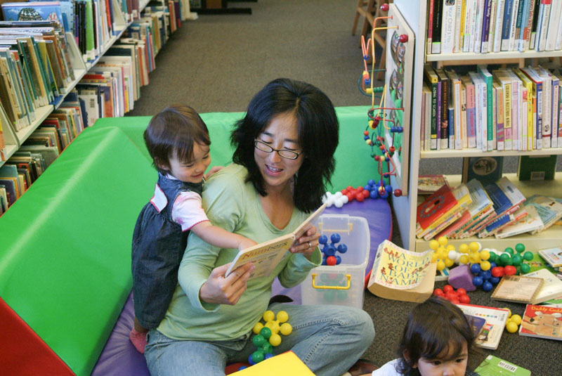 Woman sitting on colorful cushions showing a book to a toddler girl behind her.  Photo copyright Terry Lorant.