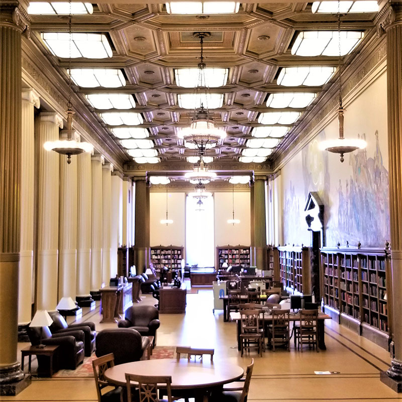 Photo of a long, tall room with bookshelves in the right wall, several tables and chairs, and ornate ceiling panels.