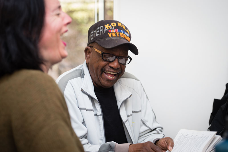 A man in a Korea veteran hat laughs while holding a book while a laughing woman sits next to him. Photo credit Becky Ruppel.