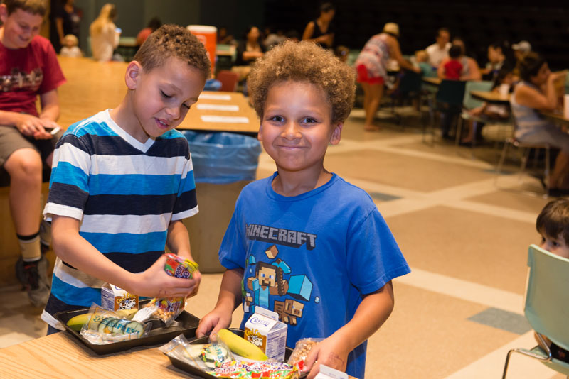 A boy in an auditorium space smiles at the camera while holding a tray with milk, banana, cucumbers, and other food. Another boy stands beside him and other children are talking, playing and eating in the background.