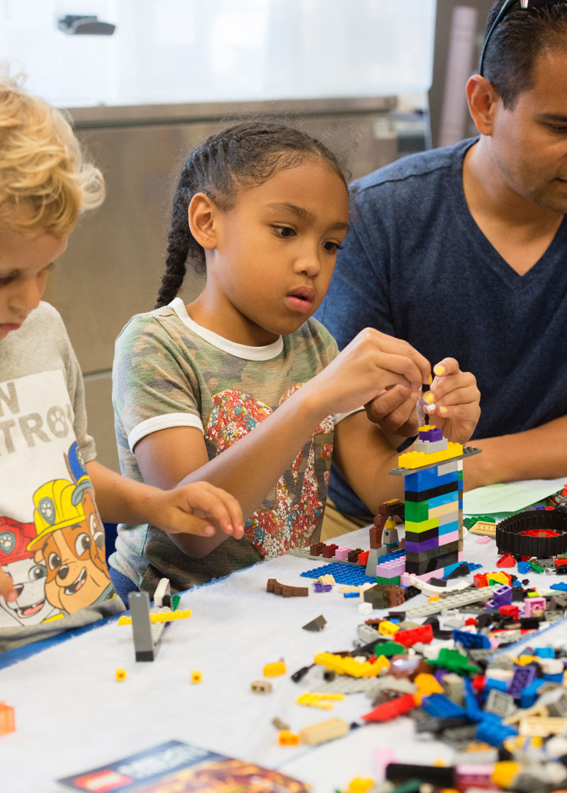 Two children and one adult sit at a table using Legos. The one in the middle is building a mutli-colored tower.