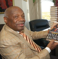 color portrait of Willie Brown; shown wearing a beige suit, seated at a table holding a copy of his autobiography. Other copies of the book are stacked on the table in front of him