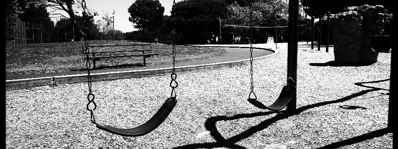 In a black and white photo, an empty park bench sites across a walking path from an empty swingset. No people are visible.