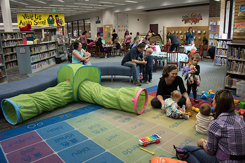 Kids and their parents sit in a play area set up inside a library. In the background are tables with activities.
