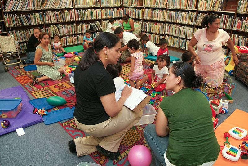 A library volunteer chats with a mother as kids play in the background.