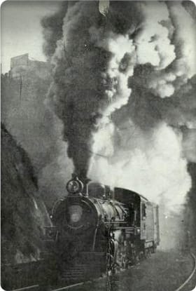 Black and white photo of a train steaming through a railway cut.