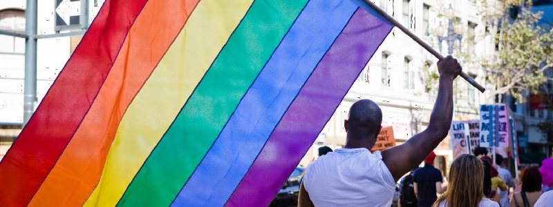 On a city street filled with people holding signs and banners, a man in a white t-shirt holds a rainbow flag in the air.