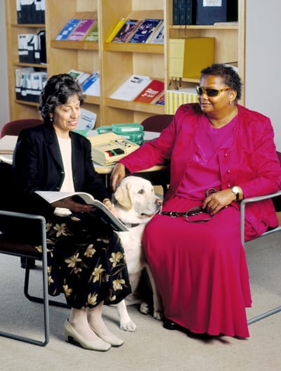 Photo of a woman reading to another woman with a guide dog at the library.