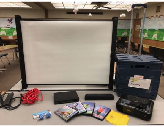 Photo of a small screen, a blue plastic box, a projector, an extension cord, a power strip, a portable speaker, a DVD player, and several DVDs.