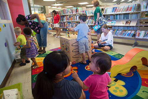 Toddlers and their guardians play on a play mat next to a long shelf of library books.