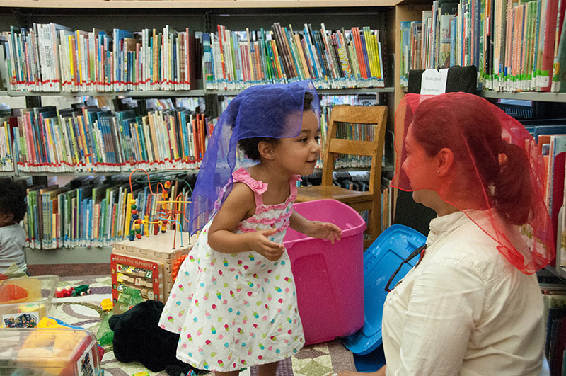 A girl and her caregiver look at each other, colorful, gauzy fabric draped over their heads.