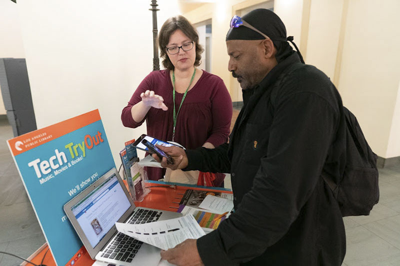 Female library staff member helping a male patron on his phone at the library’s Tech tryout station. Photo Courtesy of the Los Angeles Public Library.