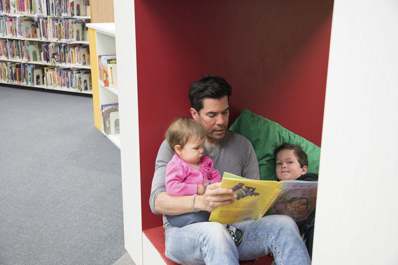 Father holding baby girl and sitting next to a young boy in a library alcove reading them a book.