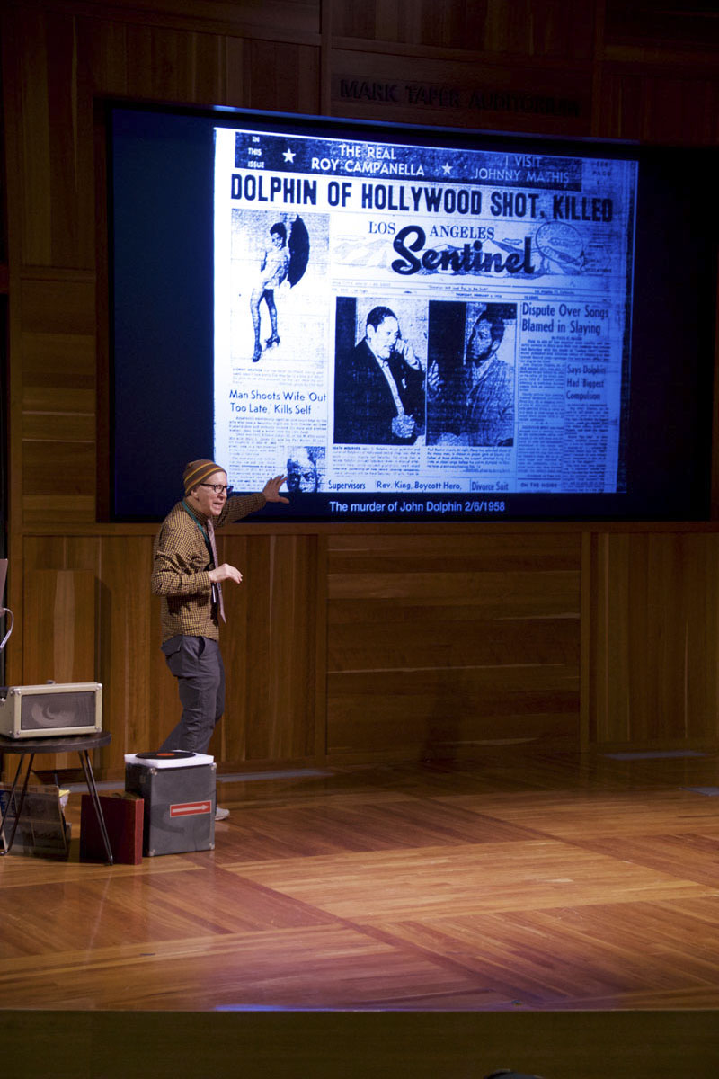 Man on-stage in an auditorium giving a presentation accompanied by a picture of a newspaper on the projection screen. Photo courtesy of the Los Angeles Public Library.