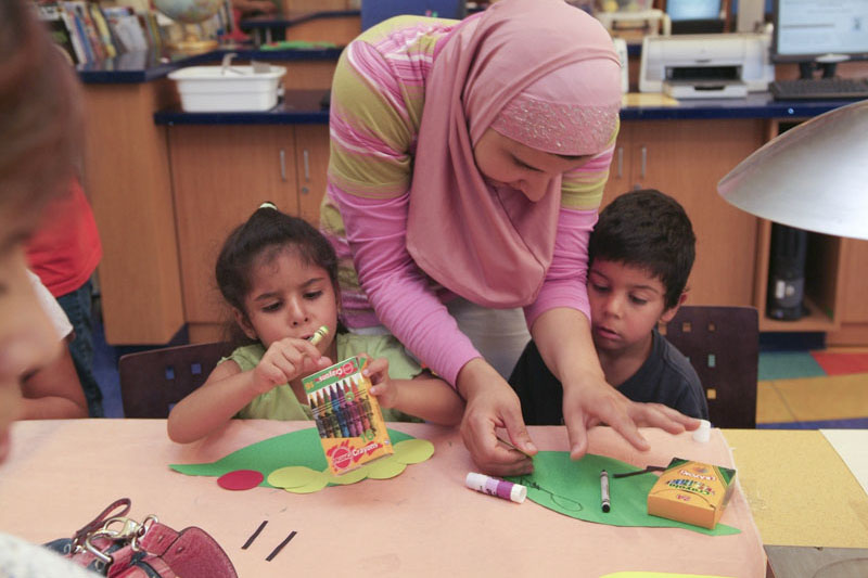 Woman leans over two young children sitting at a table and helps with and art project. Photo copyright Terry Lorant.