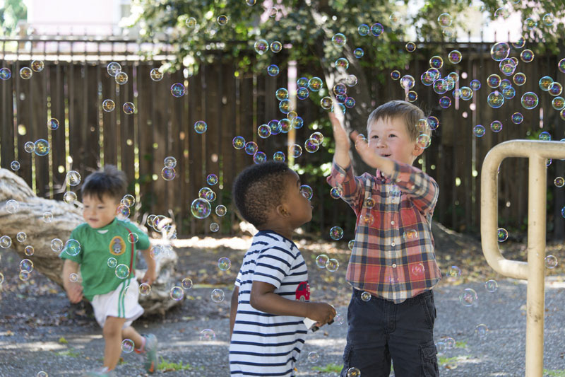 Three toddlers/young children Catching bubbles in an outside play area. Photo copyright Terry Lorant.