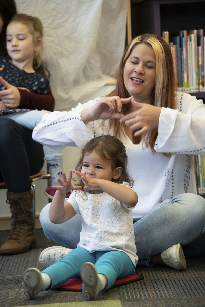 Woman and young girl sit cross legged on the ground doing hand motions to a story or song. Photo copyright Terry Lorant.