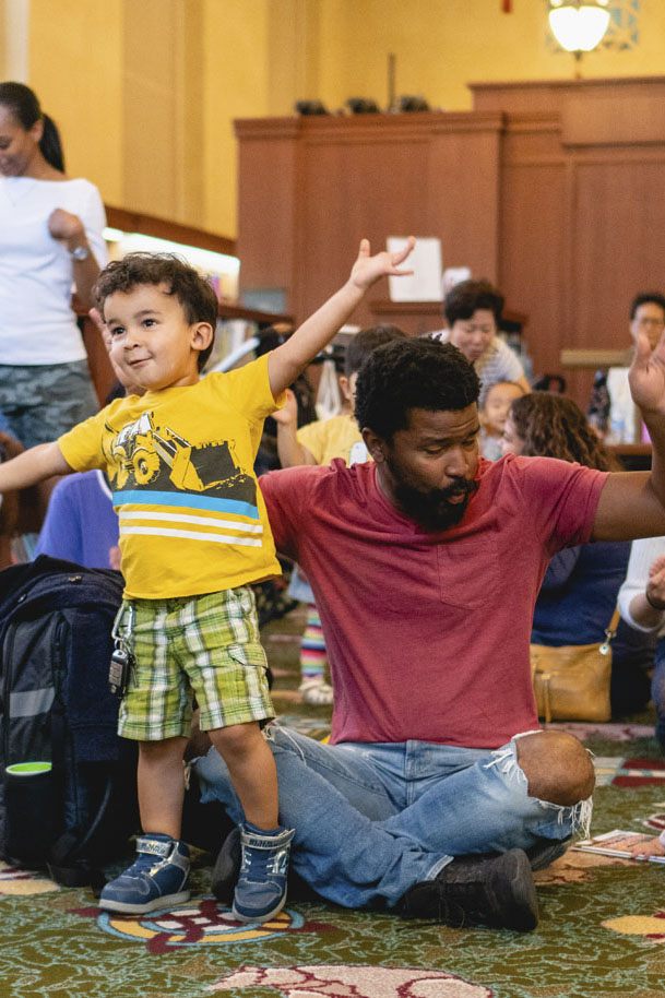 Man sitting on the floor and young child standing, taking part in storytime with other children and adults in the background. Photo courtesy Los Angeles Public Library.