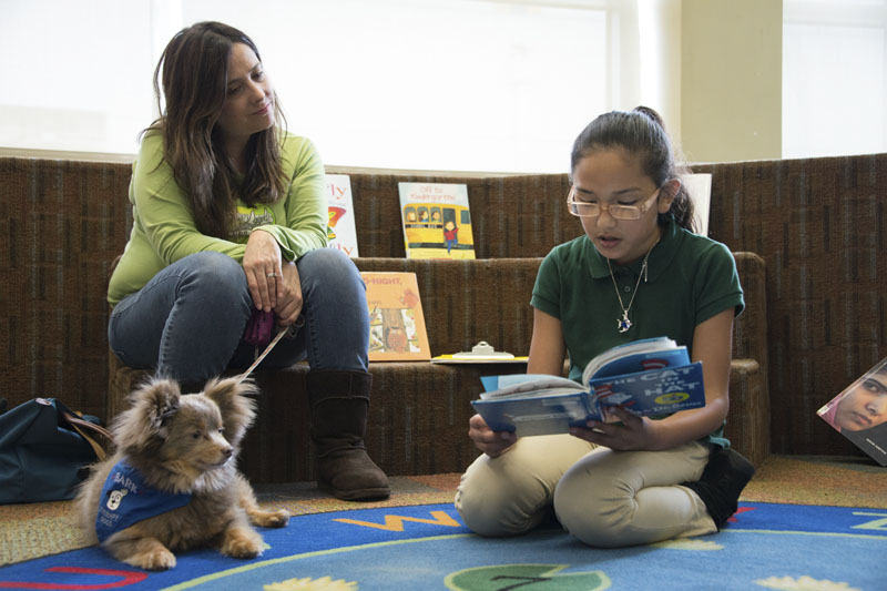 Woman sits holding a small service dog on a leash while a girl sits on the floor reading a book to the dog. Photo copyright Terry Lorant.